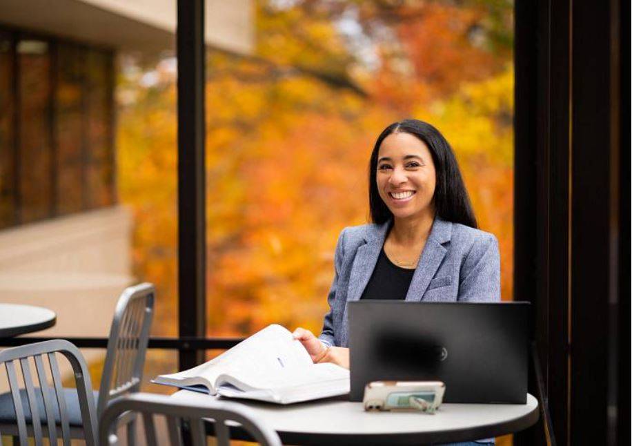 Student in grey blazer sits in front of window with textbook and laptop
